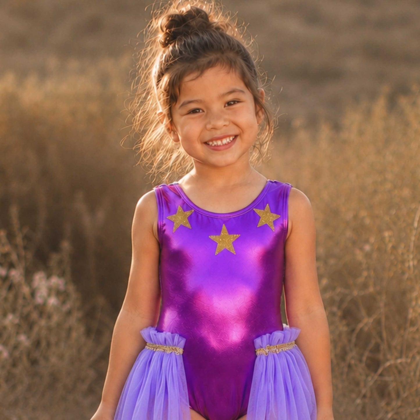 A young girl in a purple leotard with gold stars smiles at the camera, standing in a field with dry grass and wildflowers in the background.