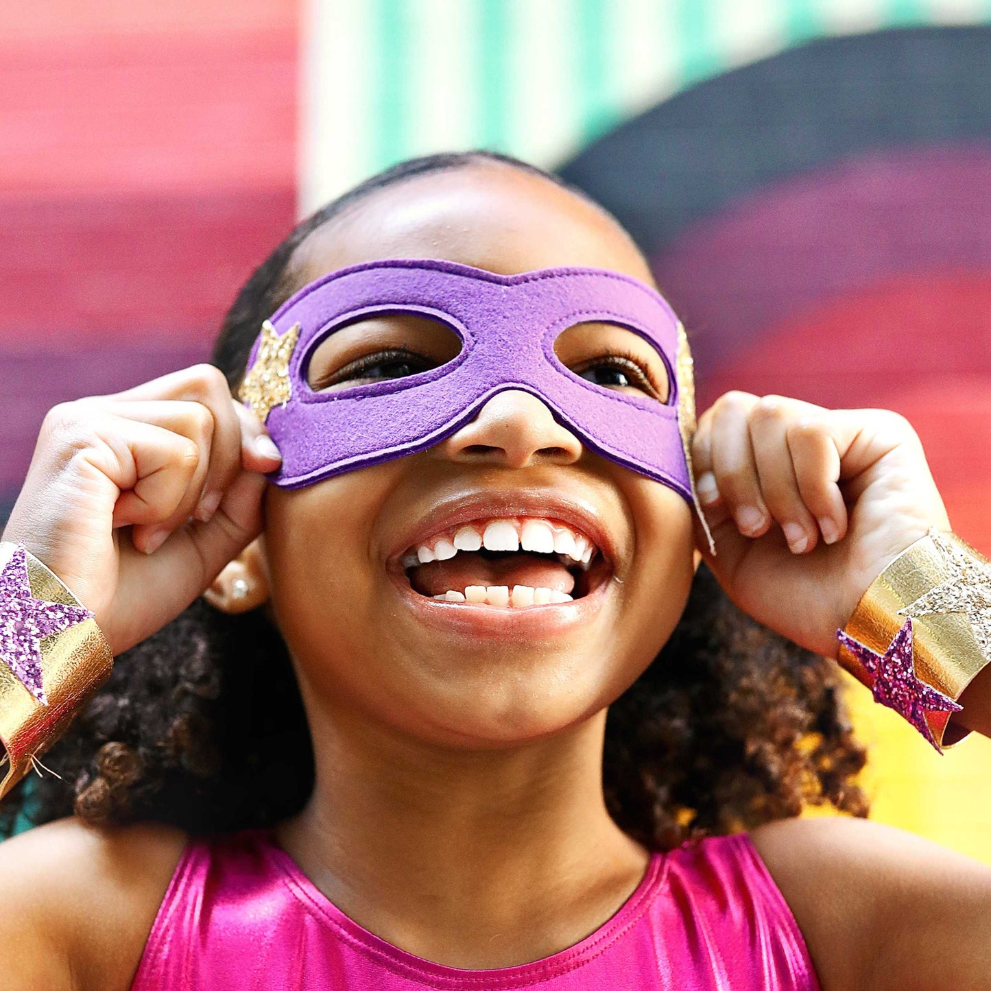 A young girl wearing a purple superhero mask and a pink top is smiling and holding her hands up to her face.