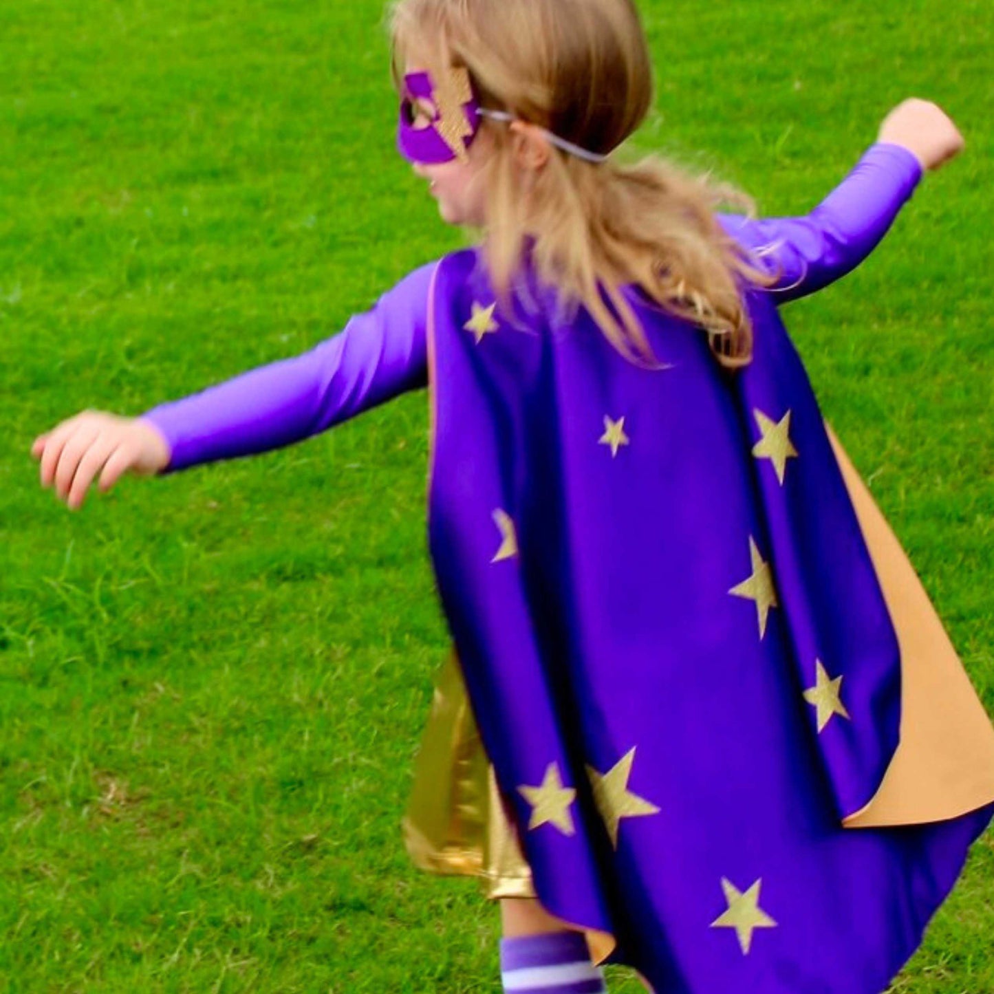A young girl in a purple cape with gold stars is running through a grassy field.