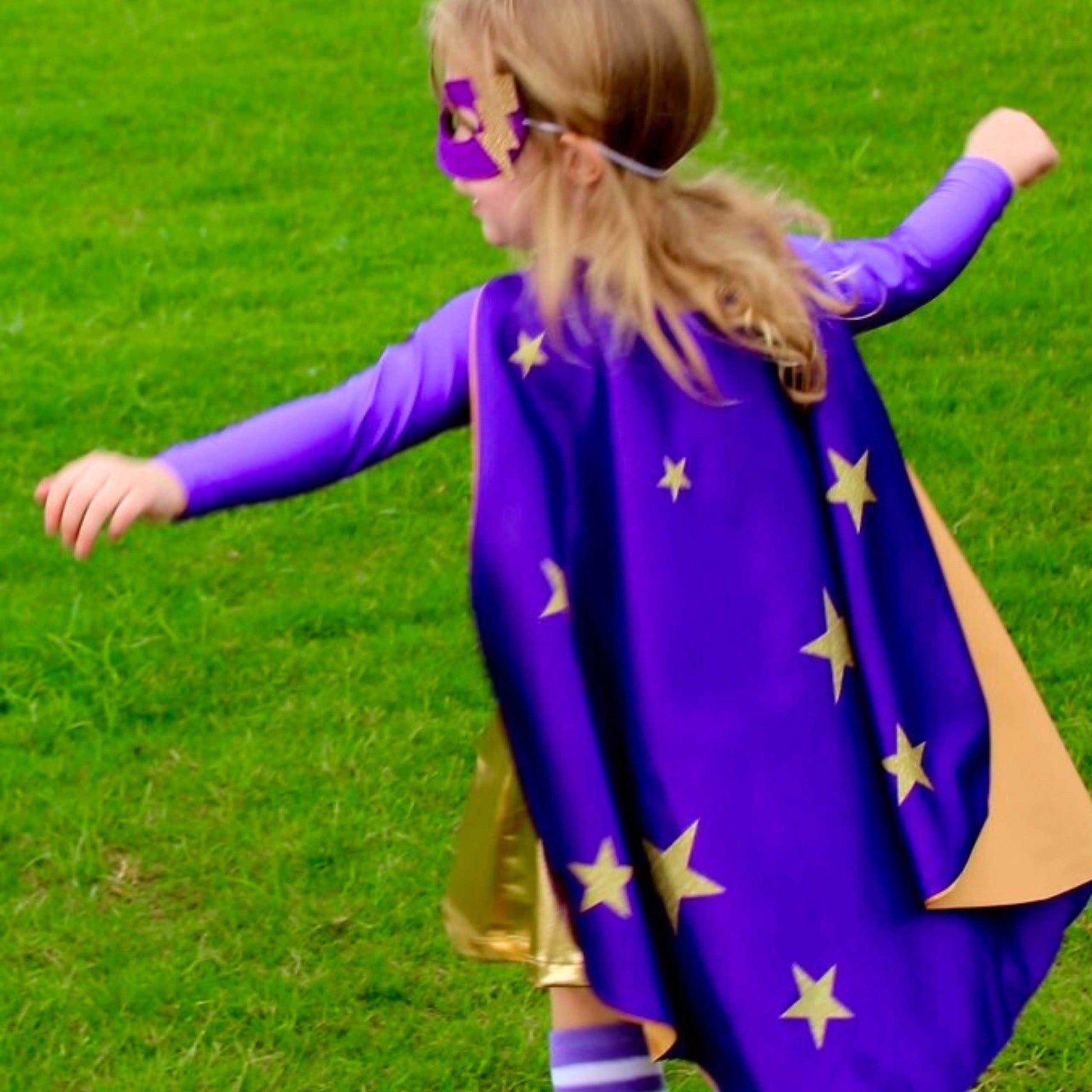 A young girl in a purple cape with gold stars is running through a grassy field.